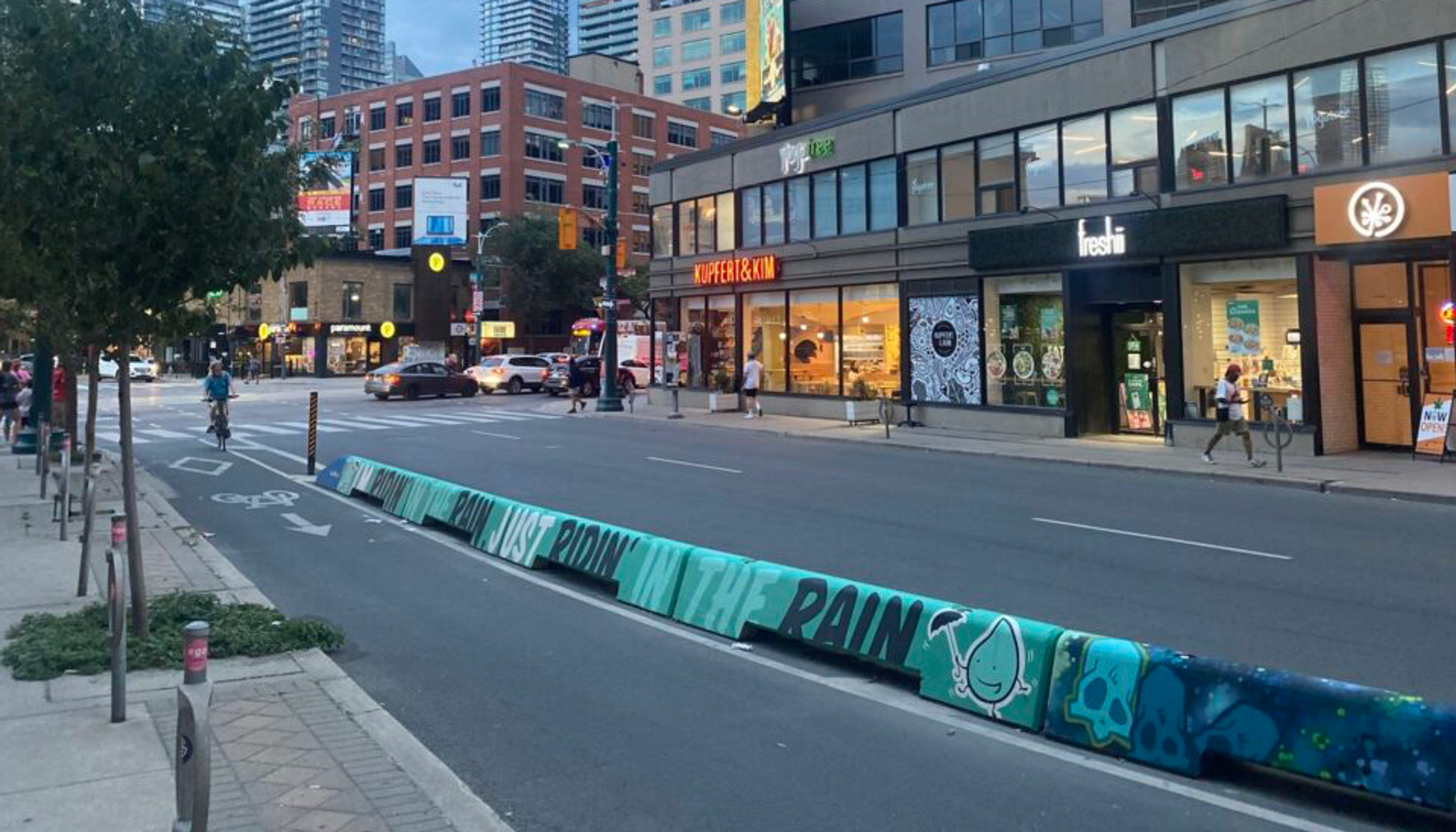 A bike lane in Toronto protected by a low concrete barrier, decorated in pastel colors with the words "Just ridin' in the rain" painted.